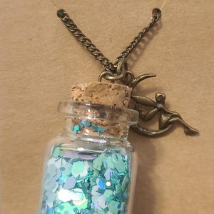 May include: A glass bottle necklace filled with iridescent blue and green glitter. The bottle has a cork stopper and is suspended from a bronze chain. A bronze fairy charm is attached to the chain.