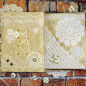 May include: A handmade journal with a brown cover decorated with white lace, crocheted doilies, and a heart-shaped design with a white ribbon and lace. The journal is closed and lying on a brick surface.