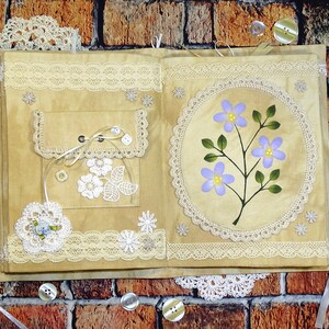 May include: A handmade journal with a beige cover decorated with lace, buttons, and a painted floral design. The journal has a pocket on the front cover and is tied closed with a ribbon.