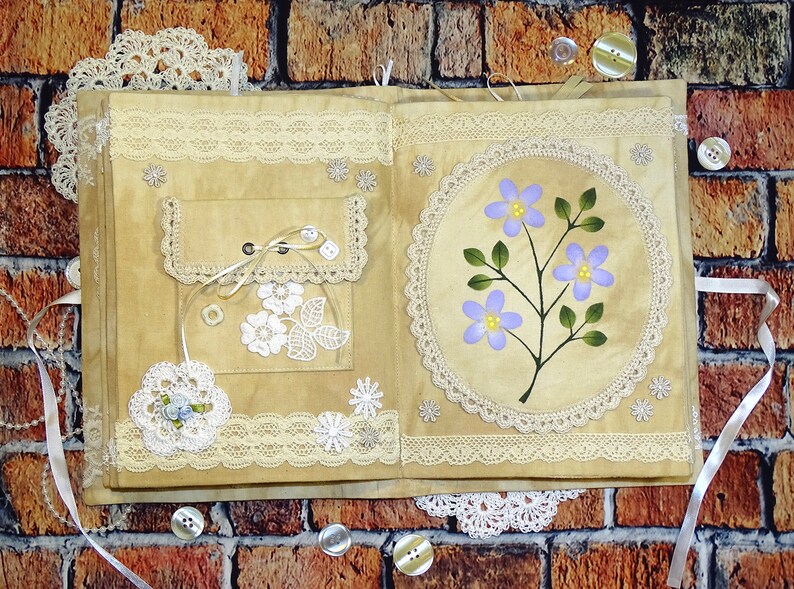 May include: A handmade journal with a beige cover decorated with lace, buttons, and a painted floral design. The journal has a pocket on the front cover and is tied closed with a ribbon.