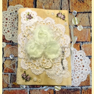 May include: A handmade journal with a rustic wood cover decorated with white lace, delicate flowers, and buttons. The journal has a vintage aesthetic and is perfect for journaling, scrapbooking, or crafting.