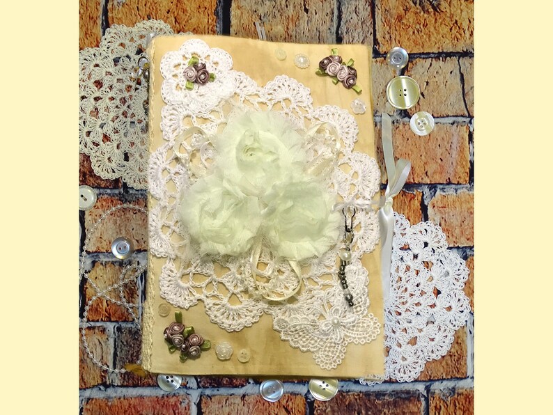 May include: A handmade journal with a rustic wood cover decorated with white lace, delicate flowers, and buttons. The journal has a vintage aesthetic and is perfect for journaling, scrapbooking, or crafting.