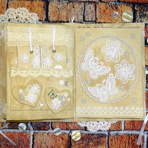 May include: A handmade scrapbook with a brown suede cover decorated with lace, buttons, and white butterflies. The scrapbook is open to reveal pages with more lace and decorative elements.
