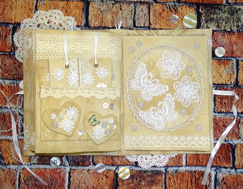May include: A handmade scrapbook with a brown suede cover decorated with lace, buttons, and white butterflies. The scrapbook is open to reveal pages with more lace and decorative elements.