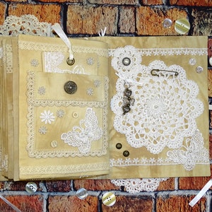 May include: A handmade journal with a beige cover decorated with lace, buttons, and a butterfly design. The journal is open to reveal a blank page.