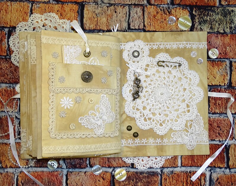 May include: A handmade journal with a beige cover decorated with lace, buttons, and a butterfly design. The journal is open to reveal a blank page.
