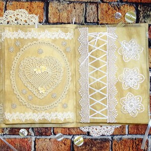 May include: A beige fabric journal with white lace trim and decorative lace appliques. The journal has a heart-shaped lace design on the front cover and a lace corset design on the back cover.