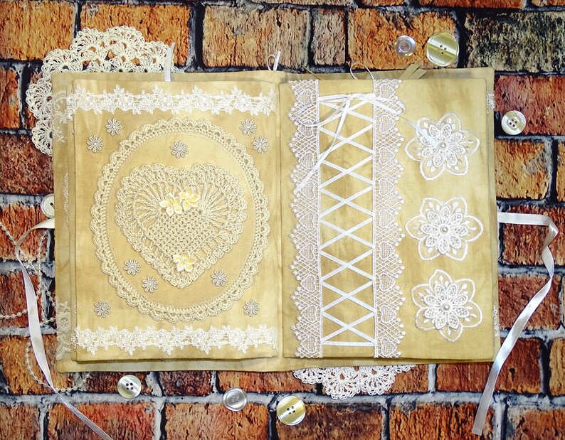 May include: A beige fabric journal with white lace trim and decorative lace appliques. The journal has a heart-shaped lace design on the front cover and a lace corset design on the back cover.