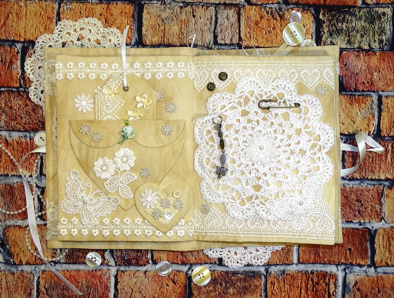 May include: A handmade journal with a brown paper cover decorated with lace, buttons, and a crocheted doily. The journal has a heart-shaped pocket on the front cover.