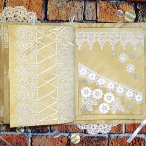 May include: A handmade journal with a brown paper cover decorated with white lace and ribbon. The journal is open to reveal pages with more lace and ribbon embellishments.