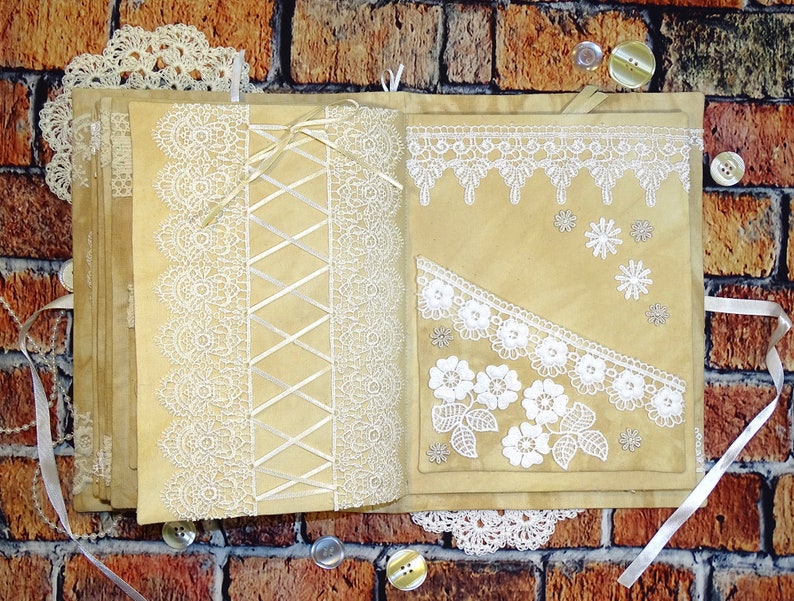 May include: A handmade journal with a brown paper cover decorated with white lace and ribbon. The journal is open to reveal pages with more lace and ribbon embellishments.