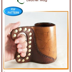 May include: A brown leather mug with a studded handle. The mug has a distressed leather finish and is made to look like a cowboy boot. The mug is being held by a hand.