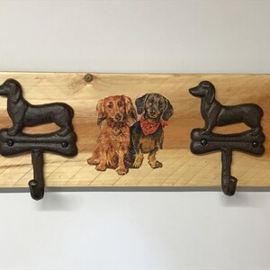May include: Wooden wall mounted coat rack with two hooks shaped like dachshunds. The hooks are black and the wood is a light brown. The rack has a printed image of three dachshunds.