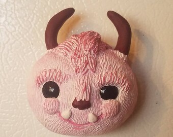 May include: A pink fuzzy creature with brown horns and black eyes. The creature has a small pink nose and a wide, toothy smile.