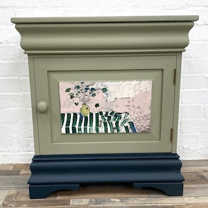 May include: A sage green bedside cabinet with a dark navy blue base and a single door. The door features a central rectangular panel painted with a still life scene of a yellow vase containing flowers on a green and white striped tablecloth.