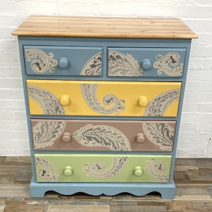 May include: A painted chest of drawers with a light wood top. The drawers are in blue, yellow, brown, and green, each with a paisley pattern. The knobs are round and match the drawer colours.