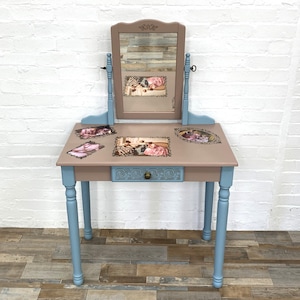 May include: A vintage dressing table with a rectangular top and a mirror. The table has a light brown top, blue legs, and a blue drawer. The mirror is framed in a matching light brown colour. Decorative floral images are on the tabletop.