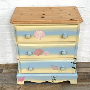 May include: A three-drawer wooden chest featuring a natural light brown wood top and a hand-painted finish with horizontal bands of pale yellow and light blue. The drawer fronts are decorated with painted pink seashells and green seaweed.