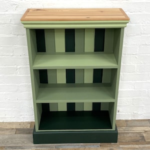 May include: A wooden bookcase featuring a natural light wood top and a frame painted in shades of sage and forest green. The back panel displays a vertical striped pattern in alternating green tones, with two interior shelves and a solid base.