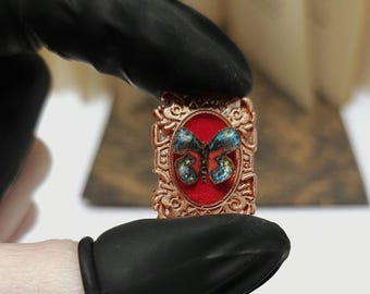 May include: A copper-toned rectangular pendant with an elaborate frame. A red backdrop features a butterfly with blue and yellow wings. The pendant is held by a person wearing a black glove.