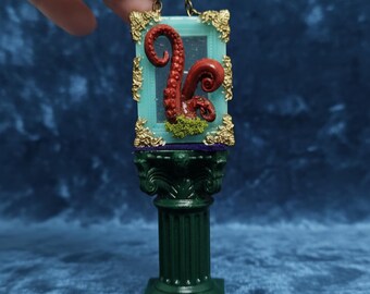 May include: Miniature sculpture of a red octopus tentacle in a teal frame with gold detailing, set on a green column base. The sculpture is suspended by a gold chain.