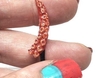 May include: A red and orange tentacle-shaped bottle stopper. The stopper has a textured surface with small, round suckers. The base is gold-coloured. The image is set against a white background.