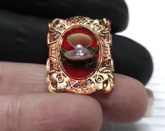 May include: A gold-coloured, ornate framed miniature scene featuring a pearl and shell on a red background. The frame has detailed scrollwork. The pearl is a light pink colour, and the shell is off-white.