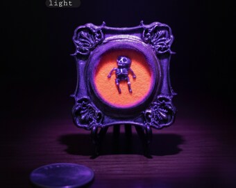 May include: A miniature framed skeleton figurine, illuminated under a blacklight, with a vibrant orange background. The ornate, dark purple frame sits on a small stand. The text in the image reads: "shown under a blacklight/uv light."
