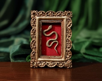 May include: A small, ornate gold-coloured frame with a detailed border. Inside, a small, detailed sculpture of a snake rests against a red background. The frame is displayed on a dark wooden surface.