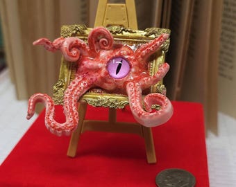 May include: A miniature sculpture of a red octopus with a large, purple eye, displayed on a small golden easel. The octopus is set within a golden frame. The display sits on a red surface, with a coin for scale.