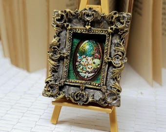 May include: Miniature framed artwork on a small wooden easel. The frame is a detailed antique gold and silver colour. The artwork features a colourful illustration with a green background. A coin is in the foreground.