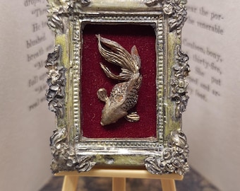 May include: A small framed artwork with a gold fish sculpture on a red backdrop. The frame has an ornate, aged look, and the piece is displayed on a miniature wooden easel. The artwork is set against the backdrop of an open book.