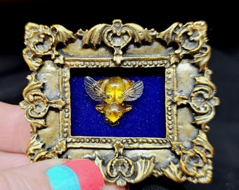 May include: An ornate, gold-coloured framed brooch. Inside, a yellow bee with transparent wings is set against a blue background. The frame has detailed scrollwork, giving it an antique appearance.