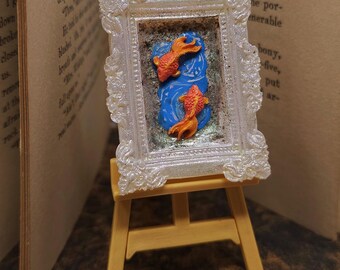 May include: A miniature artwork depicting two orange fish on a blue background, set within a white ornate frame. The framed art is displayed on a small wooden easel. A coin is placed in front.