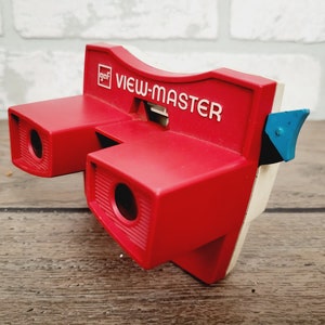May include: A red View-Master toy with a white and blue detail. The toy has two red viewing lenses and the words "View-Master" are printed in white on the top.