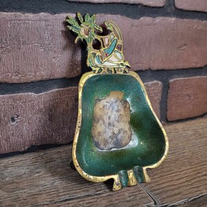 May include: A green and gold metal ashtray with a decorative design of a bird with colorful wings. The ashtray is shaped like a bell and has a small lip around the edge.
