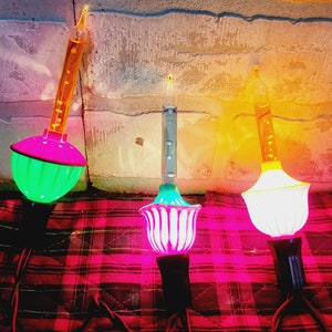 May include: Three vintage Christmas light bulbs with different colored glass bulbs and bases. The bulbs are green, white, and yellow. The bulbs are on a red and black plaid surface.