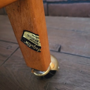 May include: Close-up of a wooden table leg with a gold-coloured caster wheel. A gold sticker on the leg reads "Genuine Rosewood, Thailand."