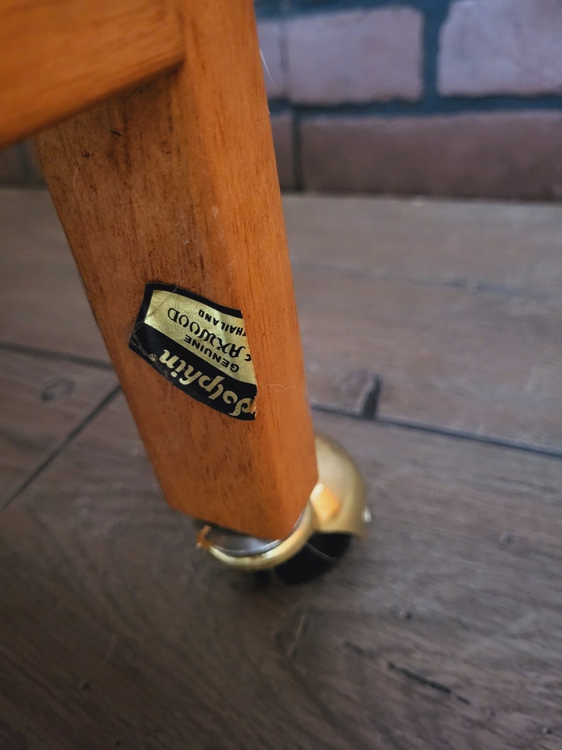 May include: Close-up of a wooden table leg with a gold-coloured caster wheel. A gold sticker on the leg reads "Genuine Rosewood, Thailand."