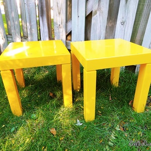 May include: Two bright yellow square end tables with four legs each, sitting on a grassy lawn.