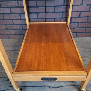 May include: A wooden serving trolley with a brown top and a lower shelf. The trolley has a black metal latch and gold-coloured wheels.