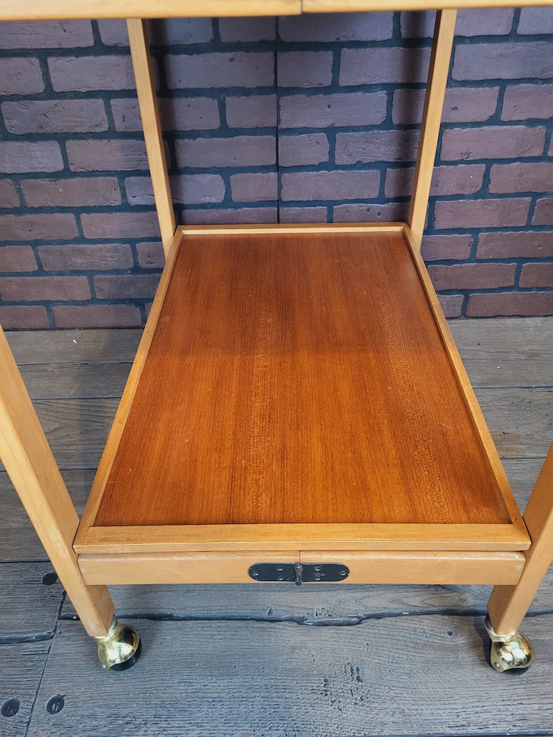 May include: A wooden serving trolley with a brown top and a lower shelf. The trolley has a black metal latch and gold-coloured wheels.