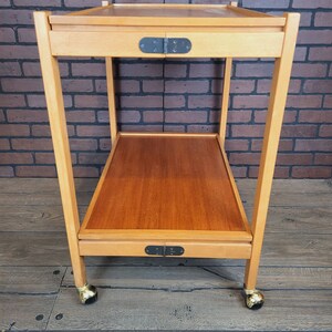 May include: A wooden serving trolley with two shelves and gold wheels. The trolley has a natural wood finish and black metal hinges.