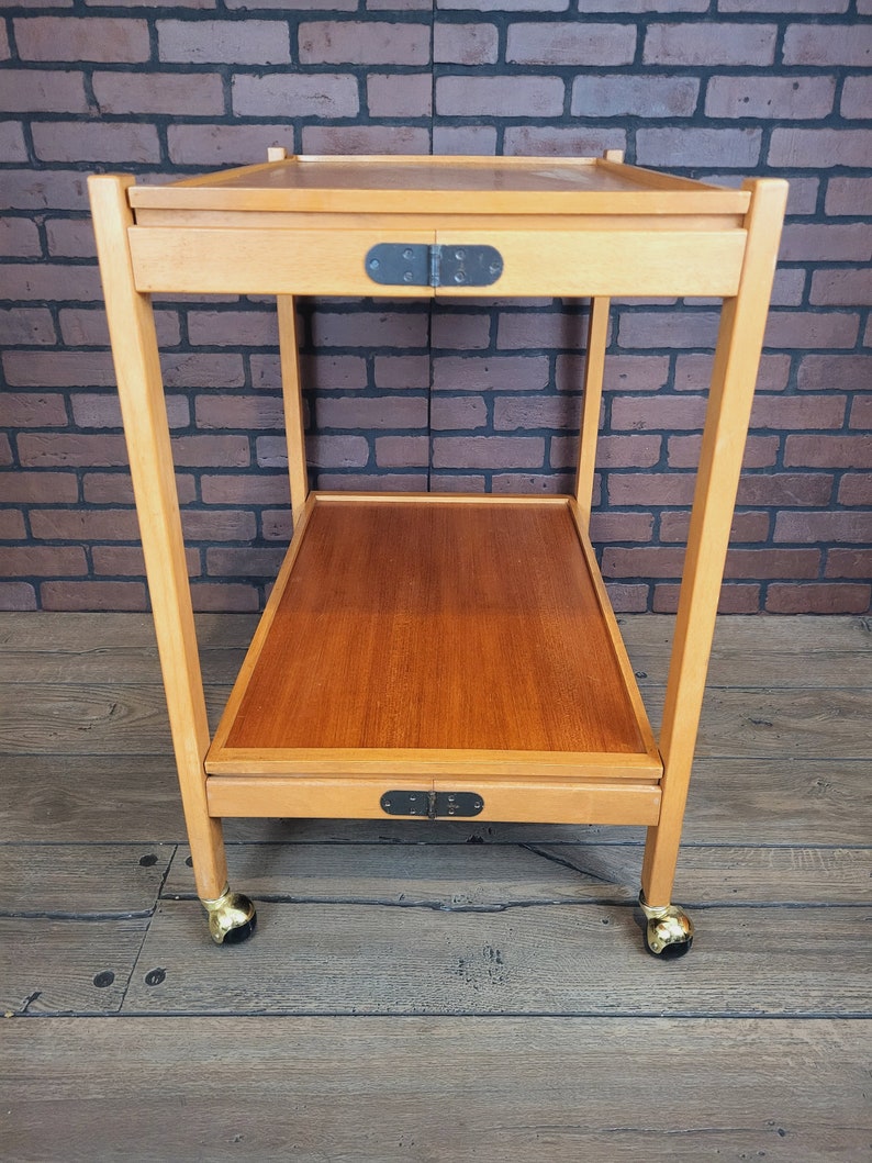 May include: A wooden serving trolley with two shelves and gold wheels. The trolley has a natural wood finish and black metal hinges.