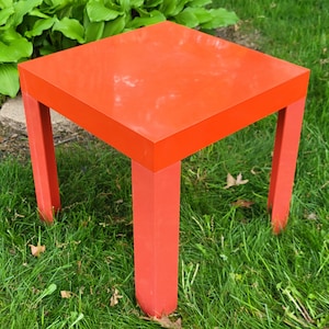May include: A bright red square end table with four legs. The table is sitting on a green grassy lawn.