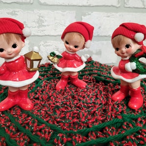 May include: Three red and white ceramic Christmas elf figurines. The elves are wearing red hats with white trim and holding Christmas-themed items. The elves are standing on a green and red crocheted surface.