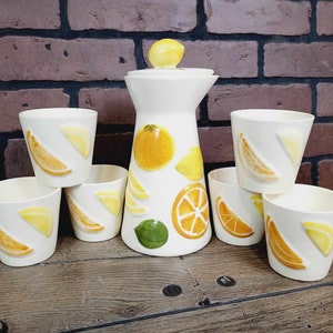 May include: A set of six white ceramic cups and a pitcher with a lemon design. The cups and pitcher are decorated with lemons, limes, and oranges.