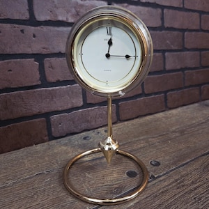 May include: A gold-toned Seiko quartz desk clock with a clear glass dome. The clock has a white face with black hands and Roman numeral hour markers. The clock is supported by a slender stand and a circular base.