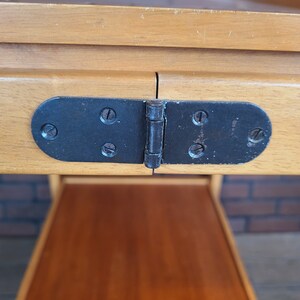 May include: Close-up of a black metal hinge with four visible screws, connecting two wooden tabletops. The hinge is attached to the underside of the table.