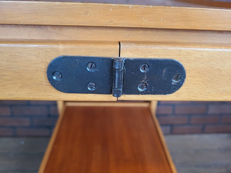 May include: Close-up of a black metal hinge with four visible screws, connecting two wooden tabletops. The hinge is attached to the underside of the table.